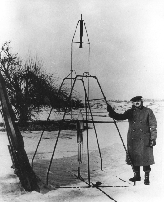 Robert H. Goddard, bundled against the cold New England weather of March 16, 1926, holds the launching frame of his most notable invention — the first liquid rocket.
