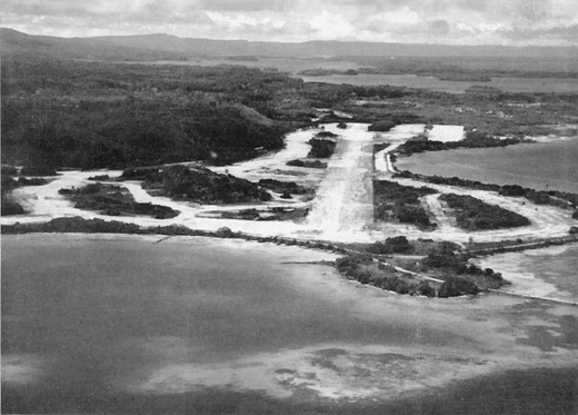 
Looking eastward, over Munda Field, toward the scene of battle, this post-war picture shows how the jungle has already begun to encroach on what was the busiest Allied airstrip in the Solomons. Within a year dense vegetation had already obscured Bibilo Hill, while once bare Kokengolo (to the center left of the photograph) sprouts a thick growth.