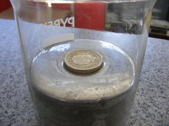 A metallic coin (an old British pound coin) floats in mercury due to the buoyancy force upon it and appears to float higher because of the surface tension of the mercury.